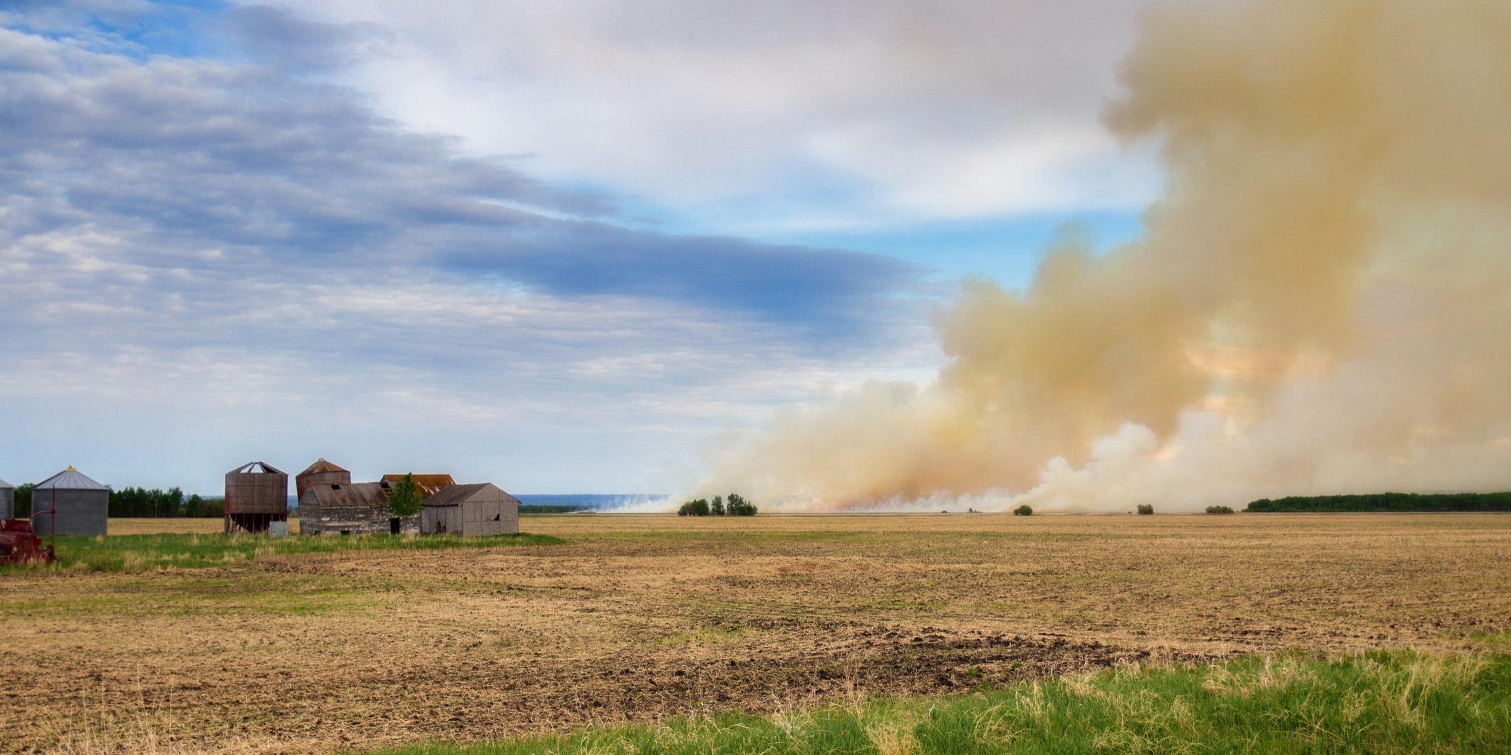 Billowing clouds of smoke from a fire burning a farm field with old sheds nearby in a summer landscape