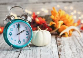 Set your clocks and fall back. Clock and decorations of mini pumpkins, colorful autumn leaves, antlers and bokeh lights over a white wooden table. Daylight saving time concept.